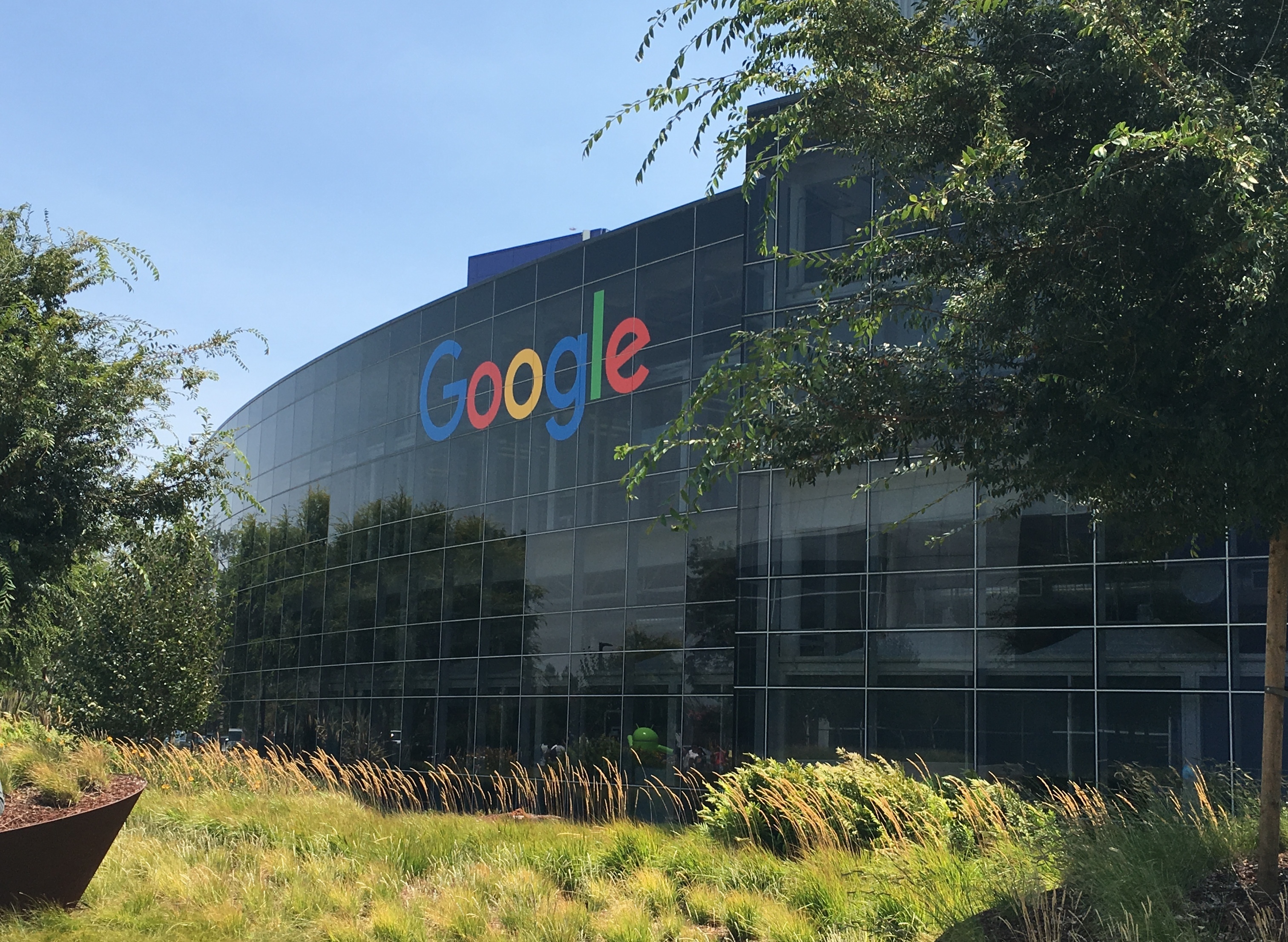 Fachada curva de vidro do Googleplex com letreiro colorido Google, árvores e céu azul.